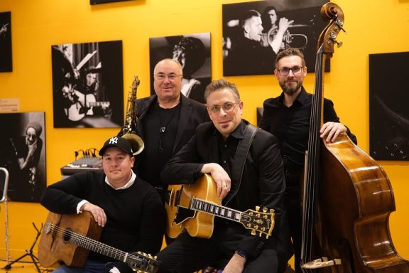 Vier Musiker posieren mit Instrumenten vor gelber Wand und Schwarz-Weiß-Fotografien.